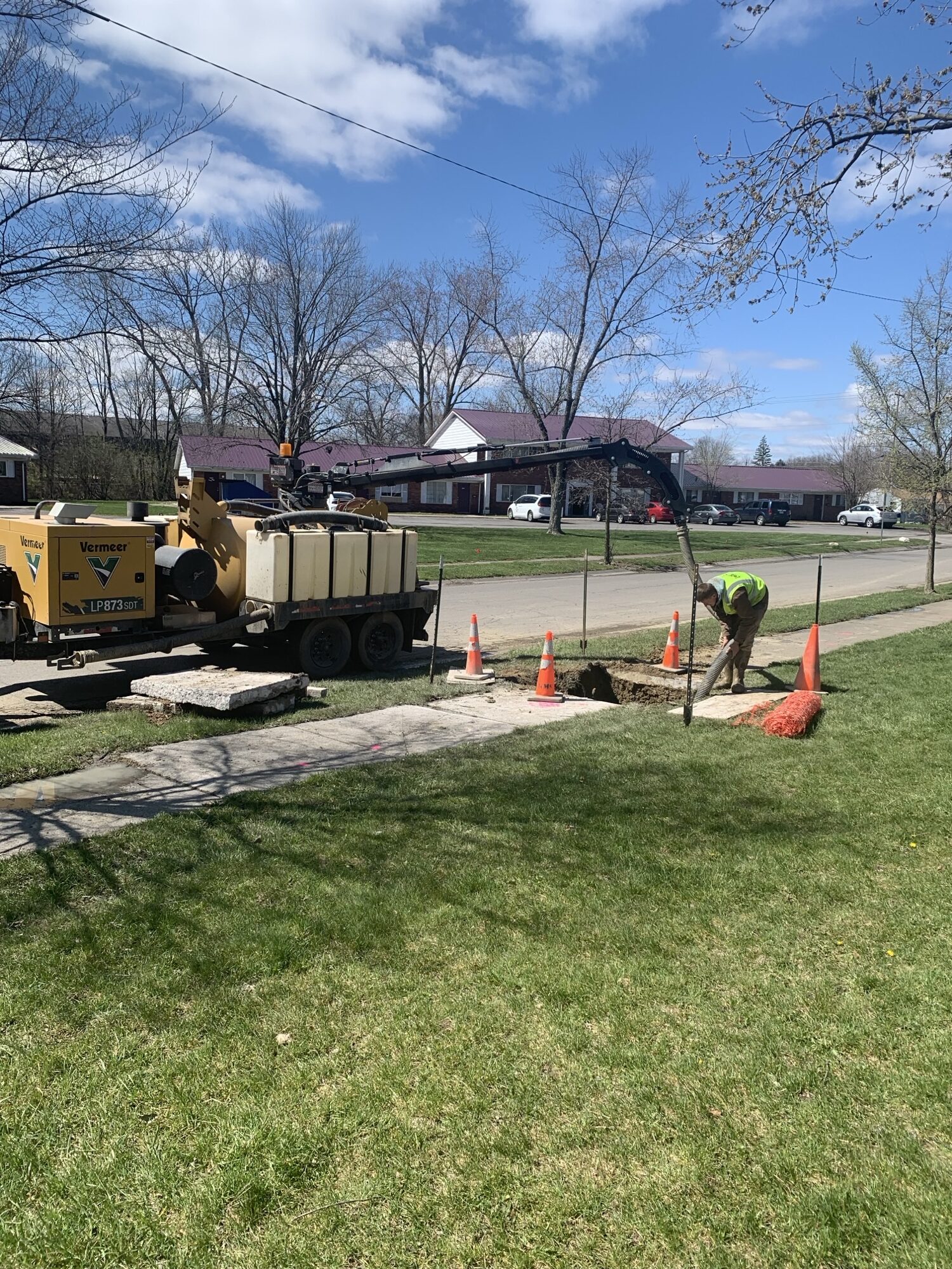 Crews are working on a water main pipe installation in a neighborhood sidewalk. Heavy machinery is being used.