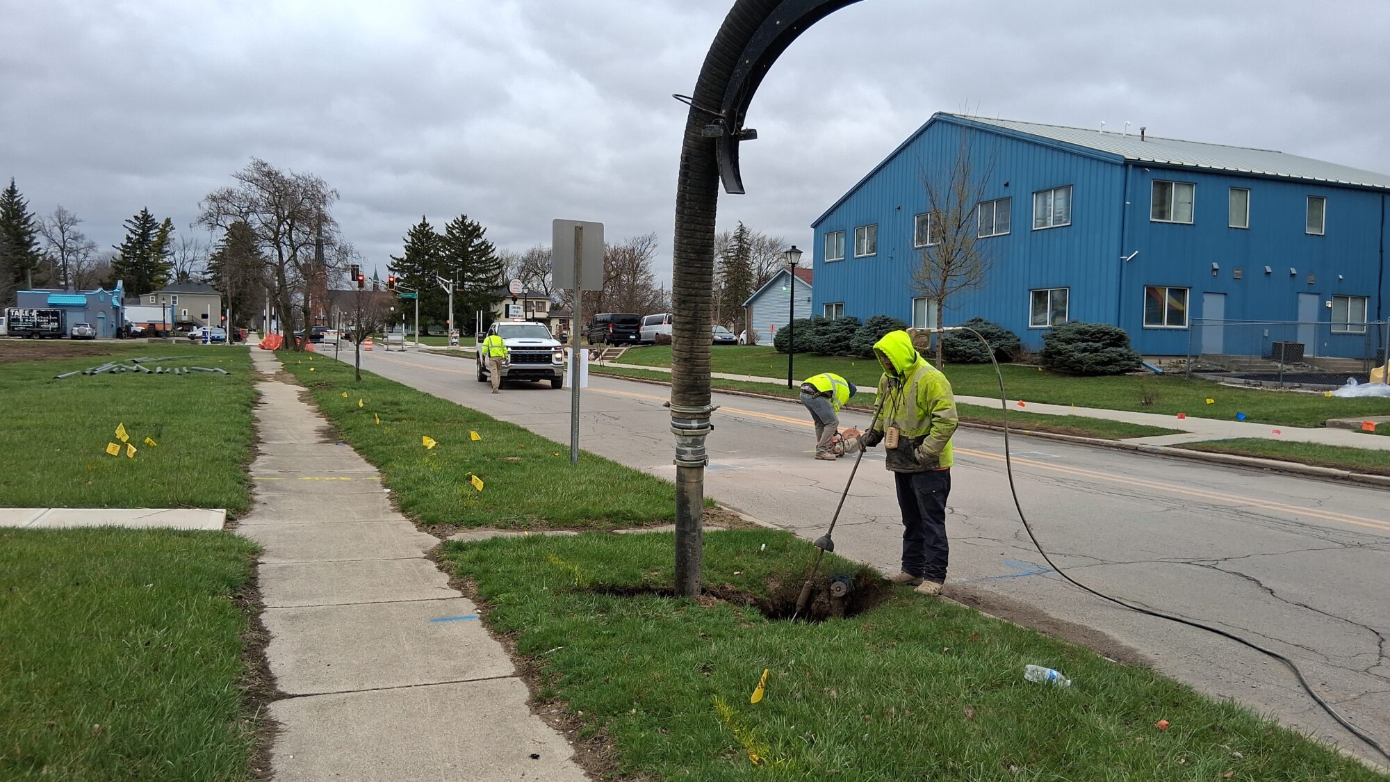 Construction crew member, wearing a yellow jacket, is seen among other workers on the street working on a pothole and setting up for a lead line replacement in a neighborhood.